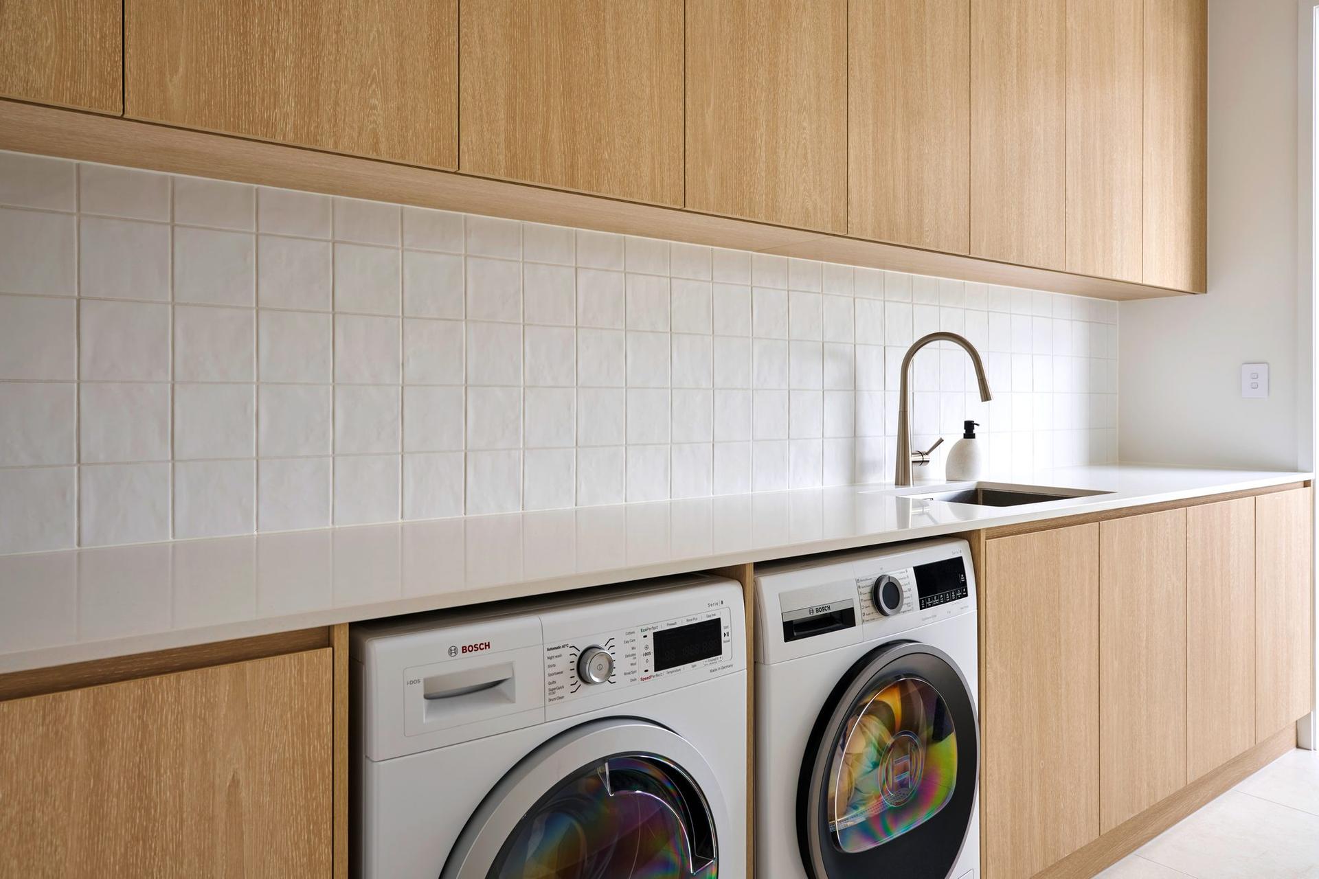 Laundry with light oak woodgrain cabinets, white tiled splashback and washing machine and dryer under the benchtop